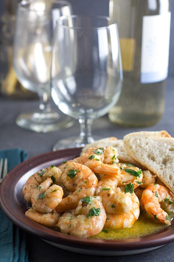 vertical image of shrimp scampi on a brown plate; wine glasses in background