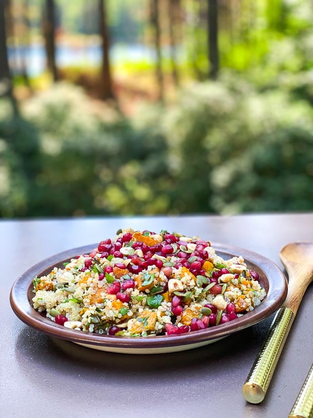 Outdoor image of roasted pumpkin and quinoa salad topped with pomegranate seeds and nuts on brown ceramic plate; wooden servers alongside