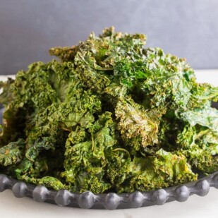 Roasted kale chips piled up on black plate, white surface, dark background