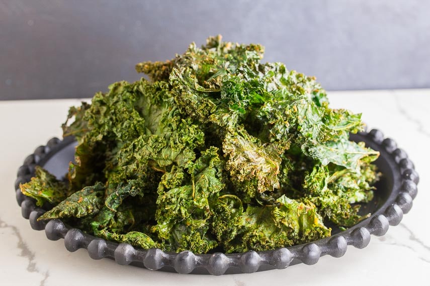 Roasted kale chips piled up on black plate, white surface, dark background
