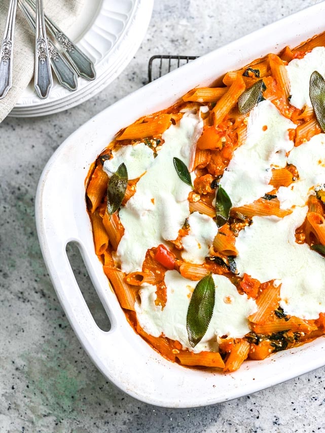 closeup image of Low FODMAP Roasted Pumpkin Baked Pasta with Sage in white oblong pan on cooling rack; white plates alongside_