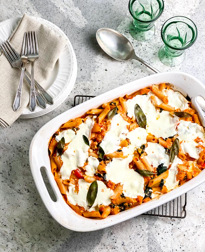 overhead image of Low FODMAP Roasted Pumpkin Baked Pasta with Sage in white oblong pan on cooling rack; white plates alongside and green glassware
