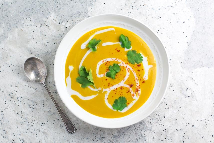 overhead image of Vegan Low FODMAP summer Squash Soup with coconut milk swirled in; white bowl and silver spoon on grey speckled quartz surface