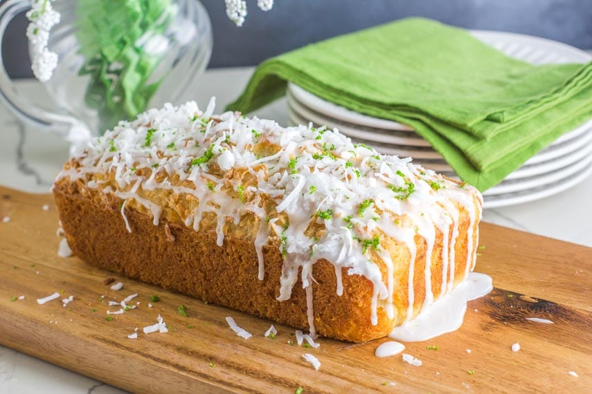 side view of coconut lime bread on a wooden board, icing dripping down; white plates and green napkin in background