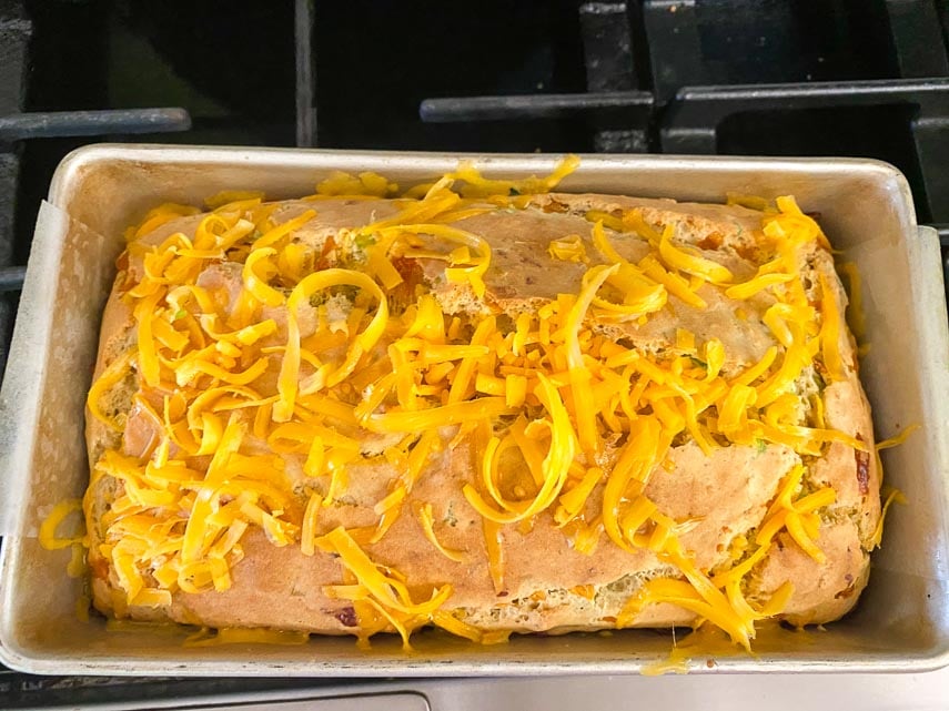 sprinkling orange cheddar cheese on top of a partially baked loaf of beer bread, sitting on stove top