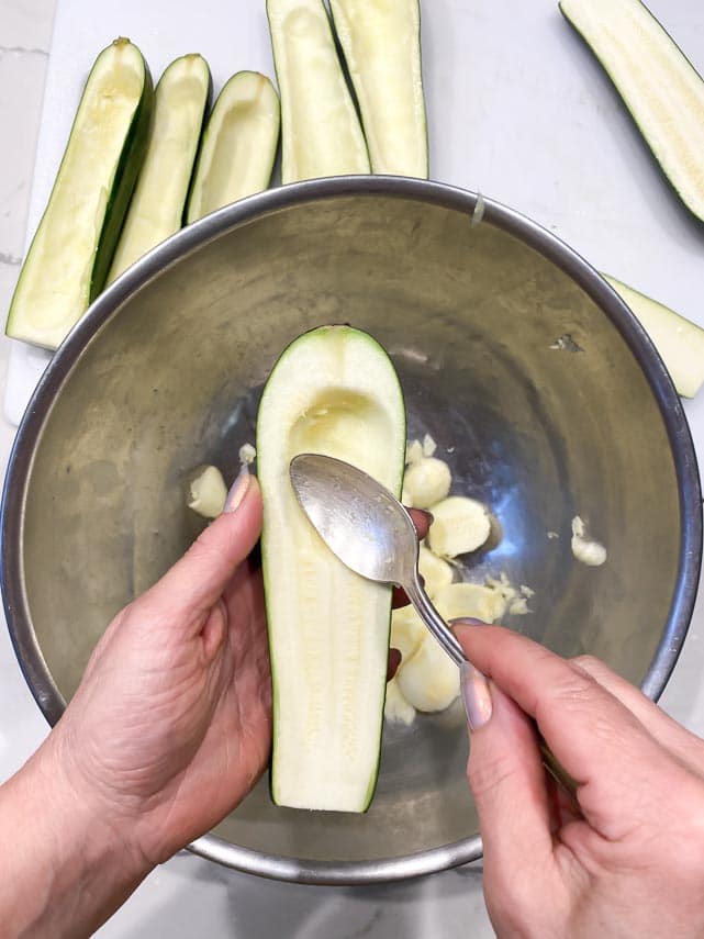 using a teaspoon to hollow out zucchini boats over a metal bowl