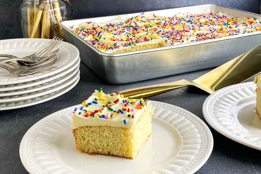 Close shot of yellow cake on white plate with vanilla frosting and rainbow sprinkles.