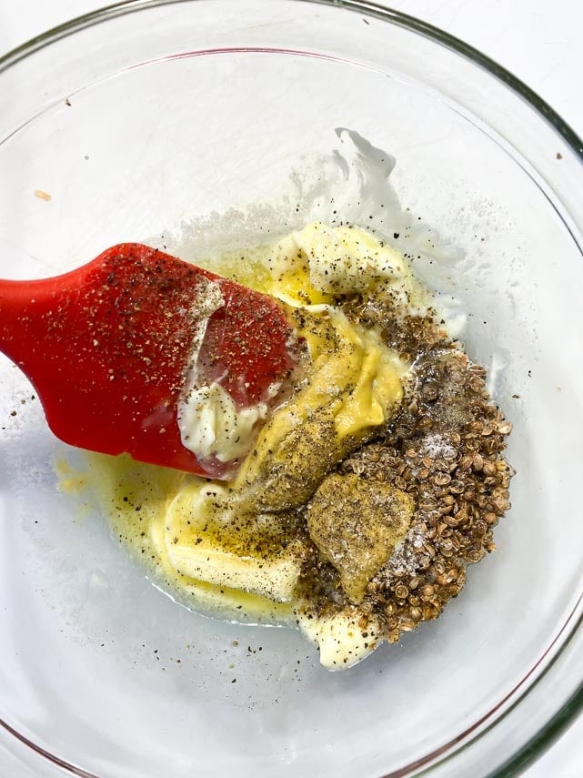 combining butter, mustard and crushed coriander seeds in glass bowl with red spatula