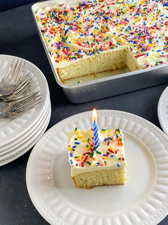 yellow-cake-with-vanilla-frosting-and-rainbow-sprinkles-on-white-plate-with-birthday-candle