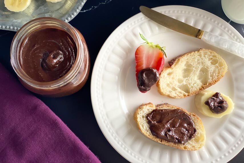Homemade Nutella in glass jar and on bread and strawberry and banana slice on white plate; dark background