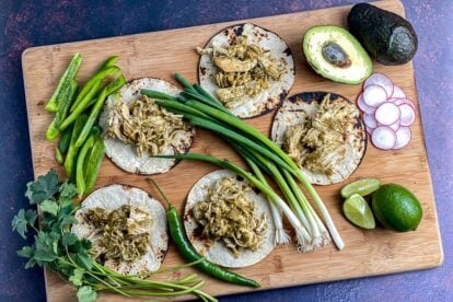overhead-view-of-Instant-Pot-Salsa-Verde-Chicken-on-corn-tortillas-on-a-wooden-board-with-scallions-avocado-hot-chile-green-peppers-radish-and-limes-1