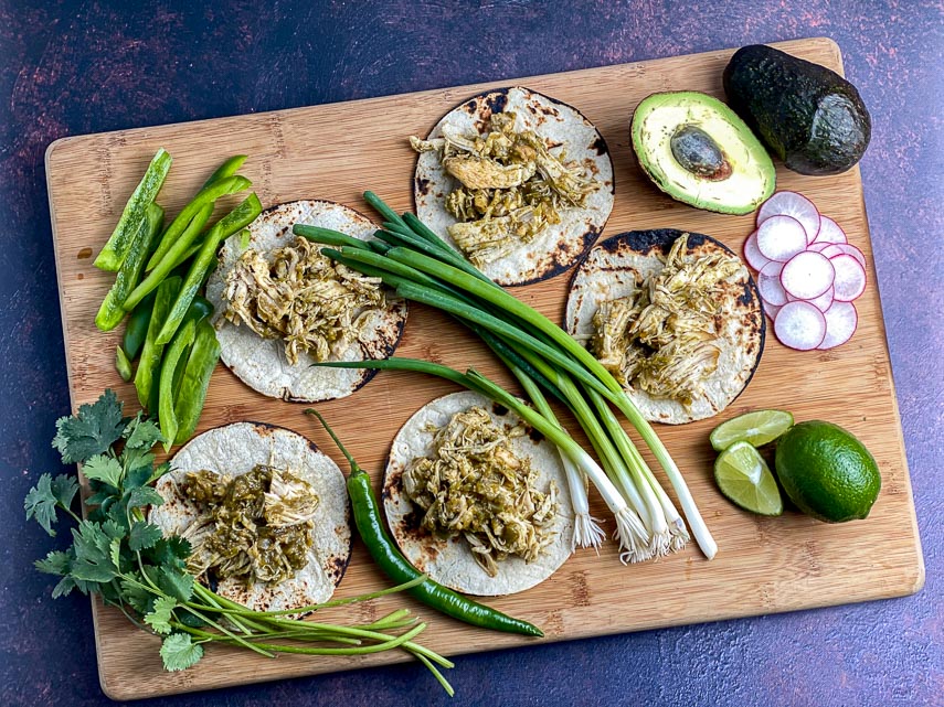 overhead-view-of-Instant-Pot-Salsa-Verde-Chicken-on-corn-tortillas-on-a-wooden-board-with-scallions-avocado-hot-chile-green-peppers-radish-and-limes-1