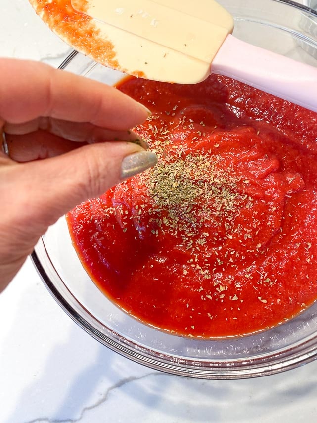 using fingers to add dry herbs to tomato purée in glass bowl