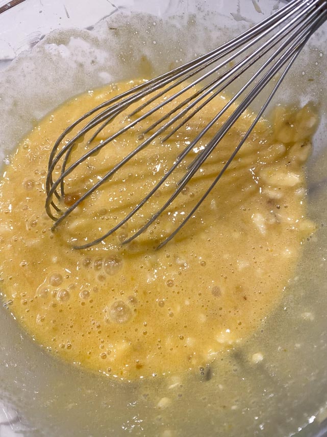 Mashing bananas with whisk in wet ingredients for banana bread in glass bowl
