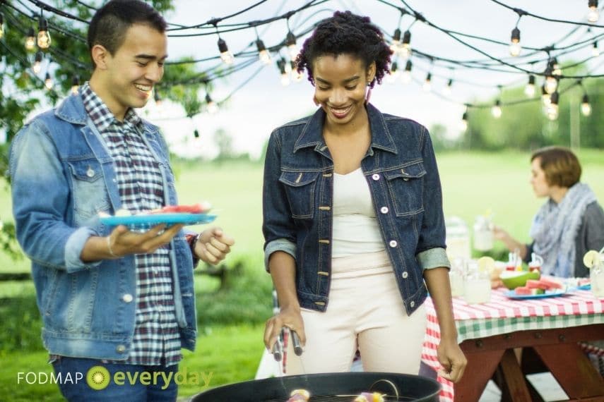 A woman grilling food on a bbq with a male friend standing next to her with a plate of food in his hand.