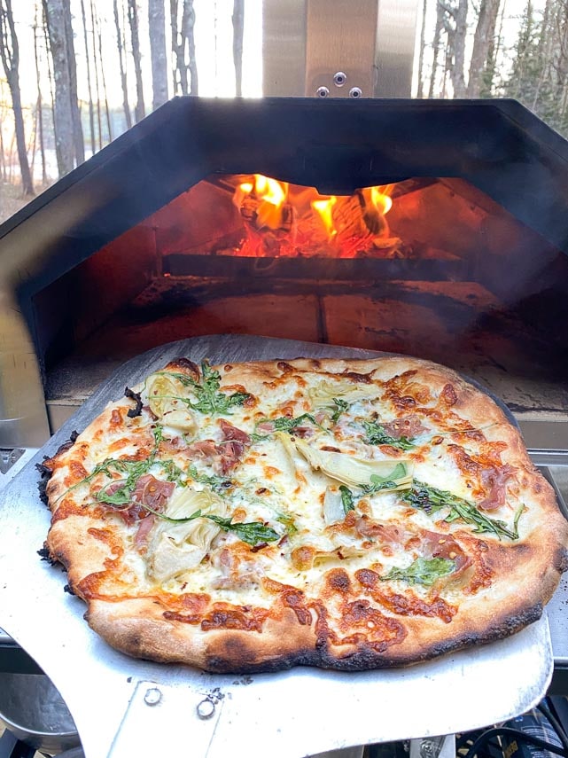 pizza with artichoke, arugula, prosciutto and cheeses in front of wood fired oven