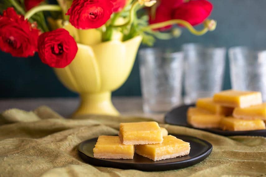 horizontal image of lemon bars on black plates; red ranunculus in vase; green napkin