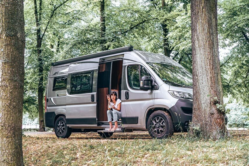 Van camping; van parked in woods with a young woman sitting in the open door flashing a peace sign. 
