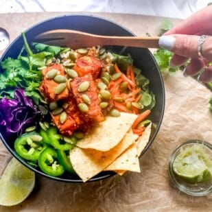 Low FODMAP Baja Tempeh Taco Salad in dark bowl; woman's hand holding wooden fork