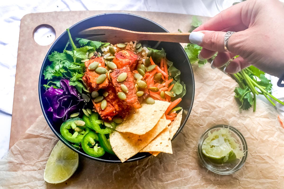 Low FODMAP Baja Tempeh Taco Salad in dark bowl; woman's hand holding wooden fork