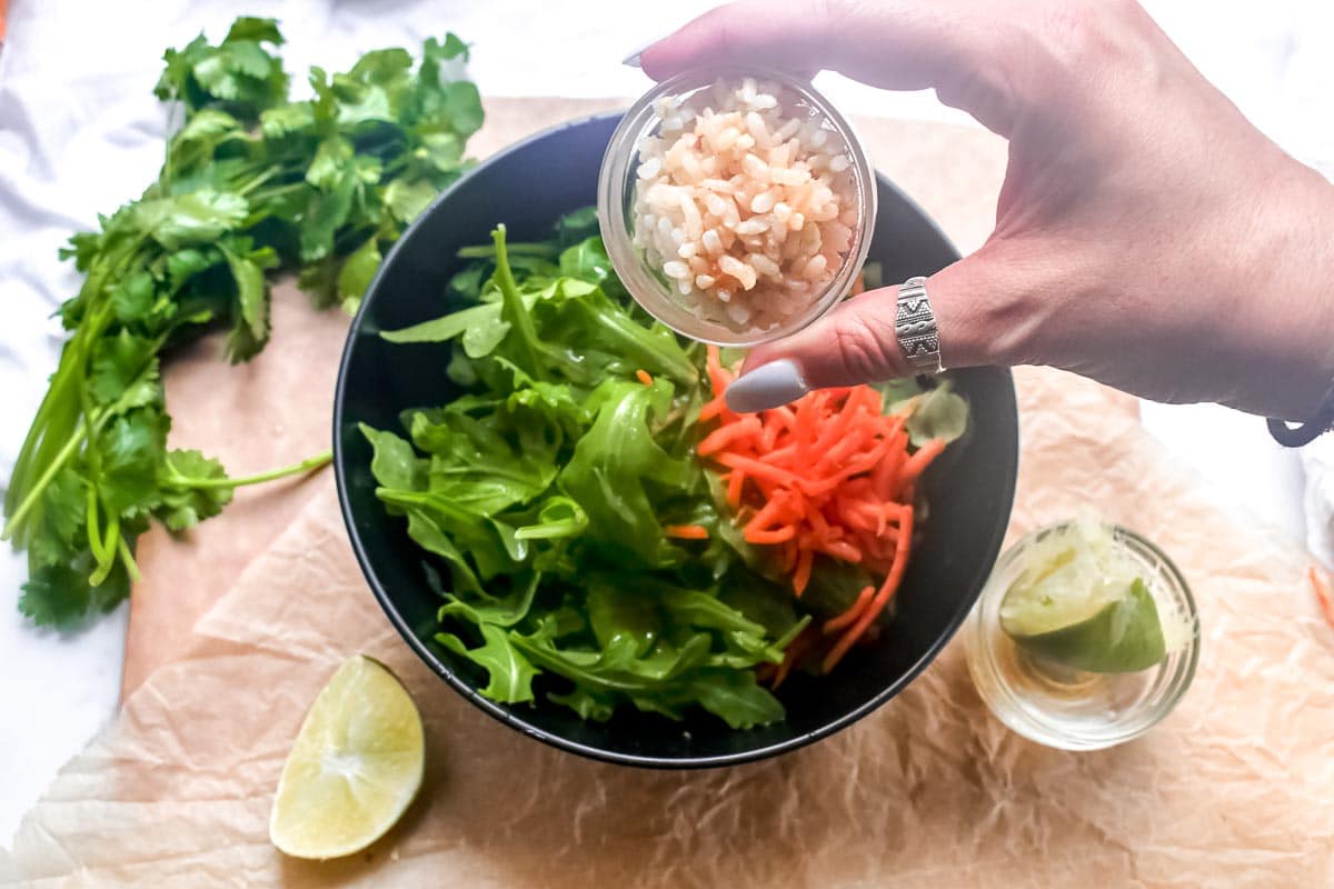 adding rice to a bowl of arugula and shredded carrots
