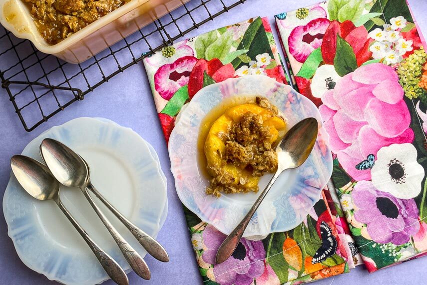 horizontal overhead image of Low FODMAP peach crisp on white plate with floral napkins and silver spoons