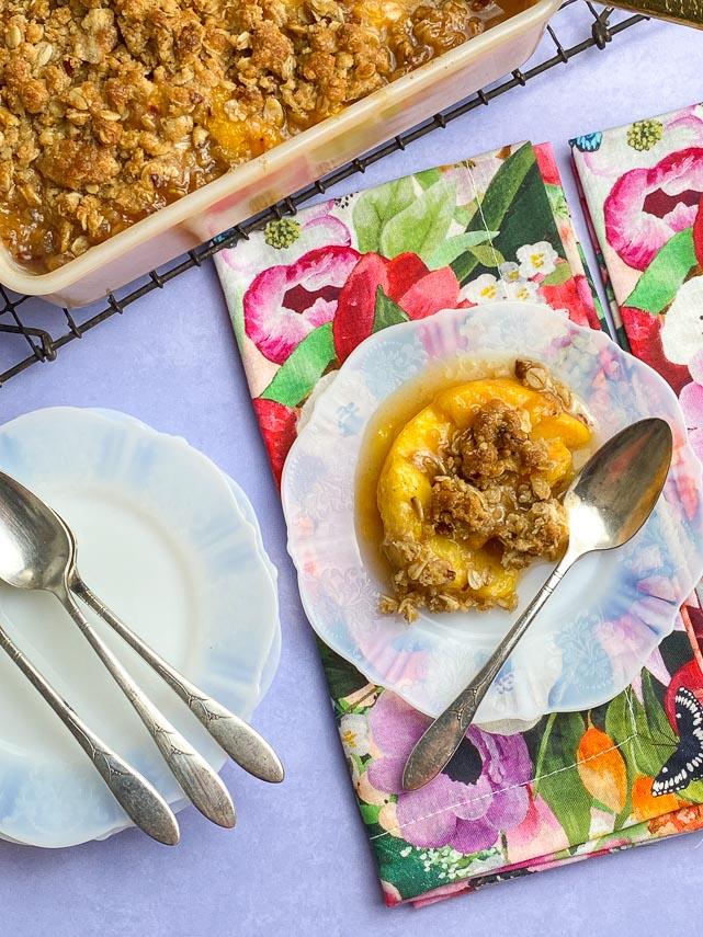 vertical overhead image of Low FODMAP peach crisp on white plate with floral napkins and silver spoons