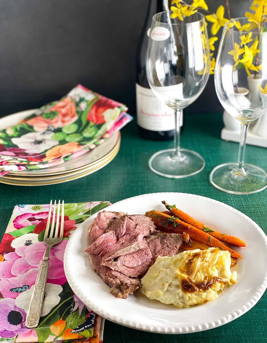 Low FODMAP Boneless Leg of Lamb, plated with scalloped potatoes and carrots on white plate; wine bottle and glasses in background