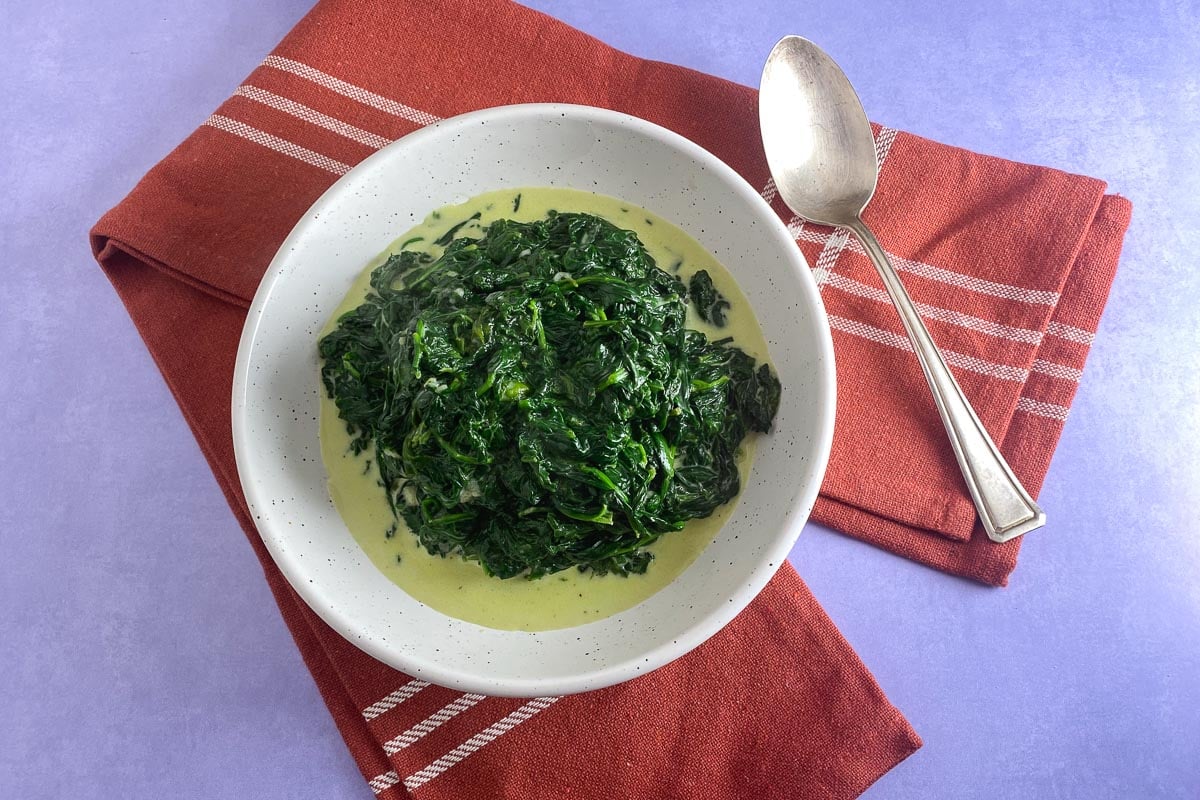 overhead image of creamed spinach in white bowl; lavender background and silver spoon