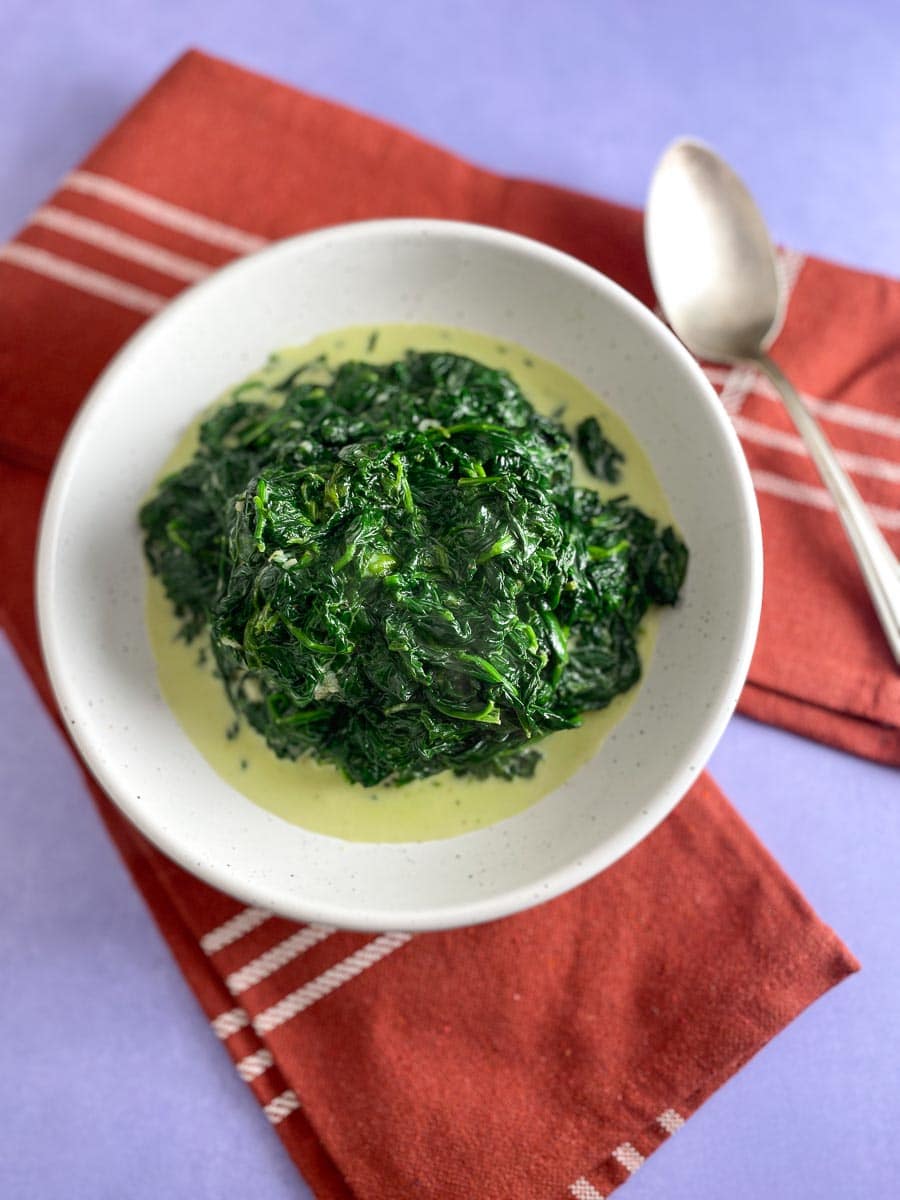 overhead vertical of creamed spinach in white bowl; lavender background and silver spoon