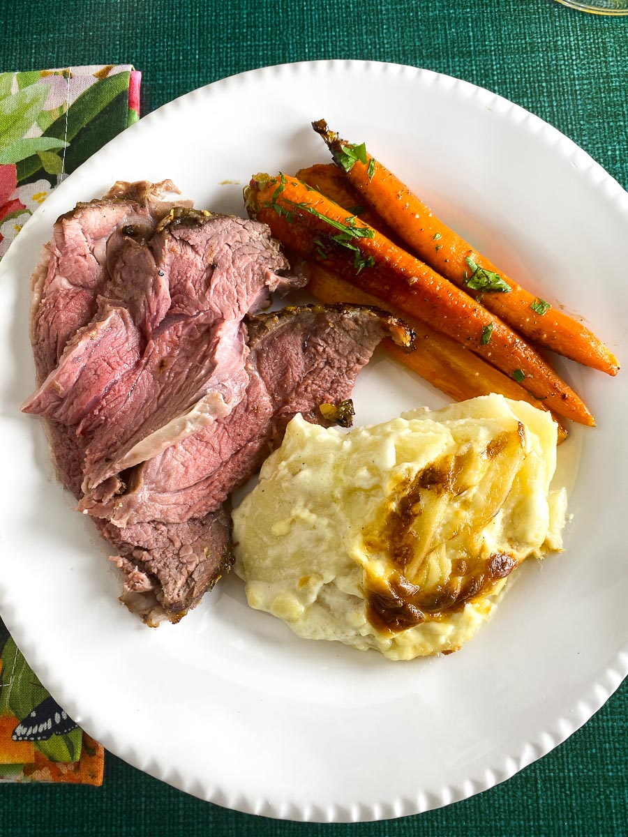 vertical image of Low FODMAP Boneless Leg of Lamb, plated with scalloped potatoes and carrots on white plate; flowered napkin; green tabletop surface