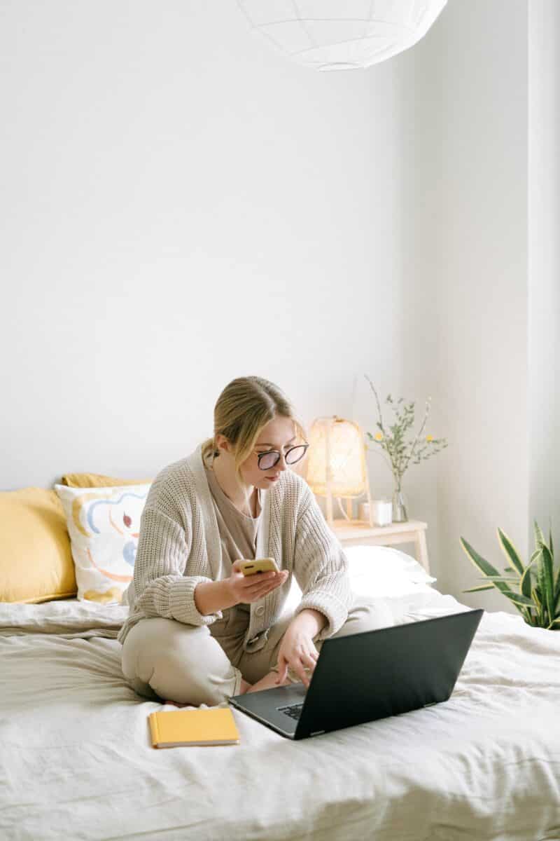 blonde woman on bed looking at smartphone and computer