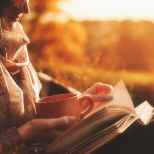Beautiful girl in autumn forest reading a book covered with a warm blanket.a woman sits near a tree in an autumn forest and holds a book and a cup with a hot drink in her hands. Girl reading a book.