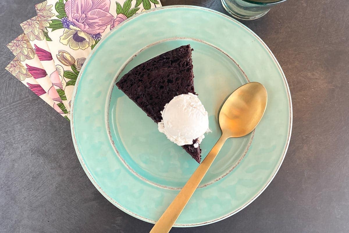 wedge of Gluten free Low FODMAP Maple Mocha Cake on aqua plate with vanilla ice cream melting on top; water glass and gold spoon, paper napkins