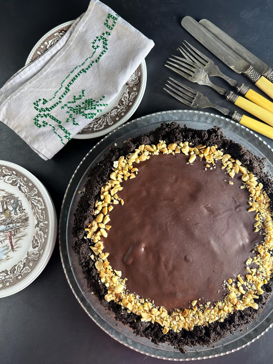 vertical of Low FODMAP Chocolate Peanut Butter Pie, dark background; antique plates, silverware and embroidered napkin
