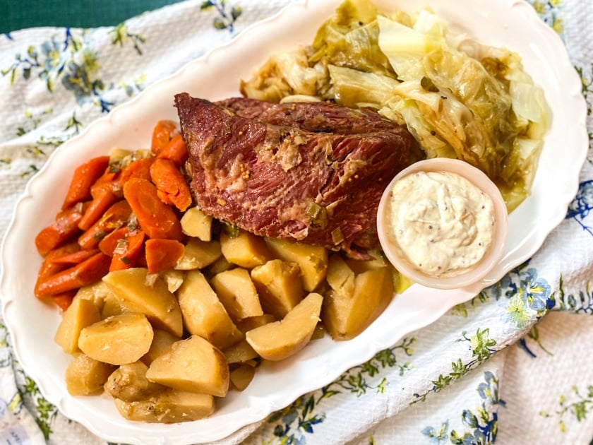 overhead image of Low FODMAP Slow Cooker Corned Beef on oval white platter with cabbage, carrots and potatoes - green backdrop