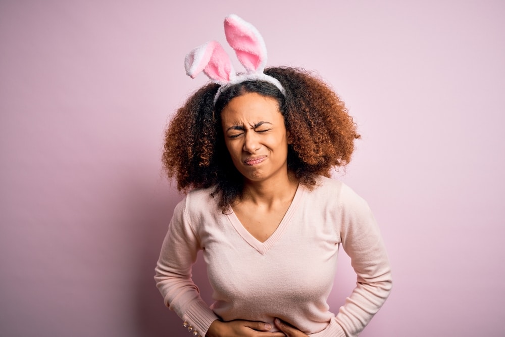 Young african american woman with afro hair wearing bunny ears over pink background with hand on stomach because indigestion, painful illness feeling unwell. Ache concept.