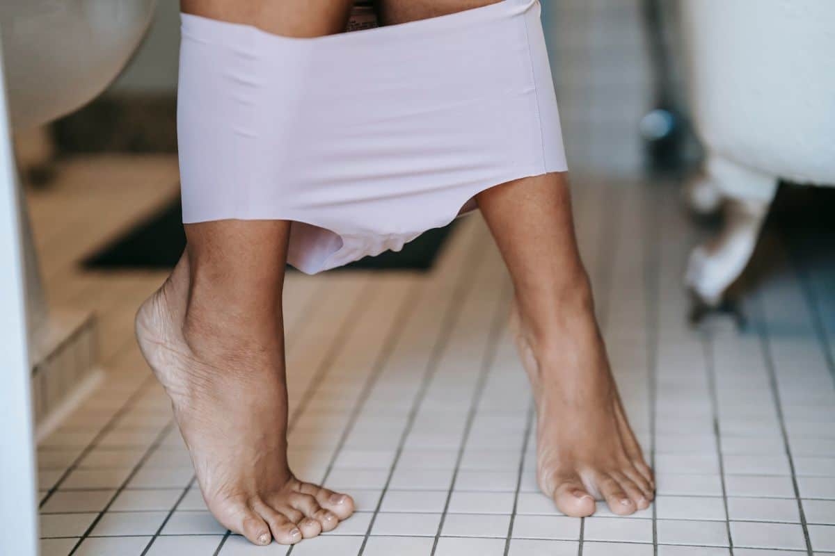 side view of a woman's bare feet with her underpants around her ankles as she sits upon a toilet in a tiled bathroom