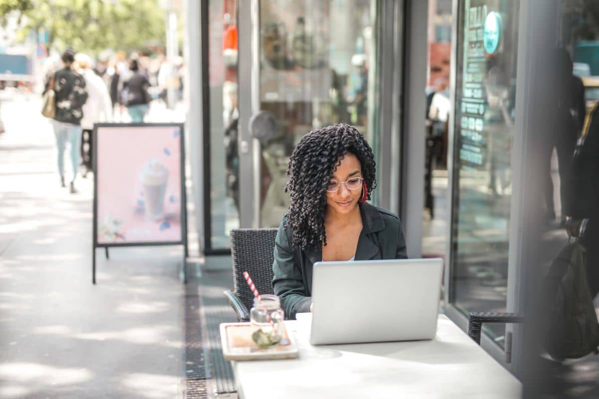 Ethnic-young-woman-using-laptop-while-having-tasty-beverage-in-modern-street-cafe