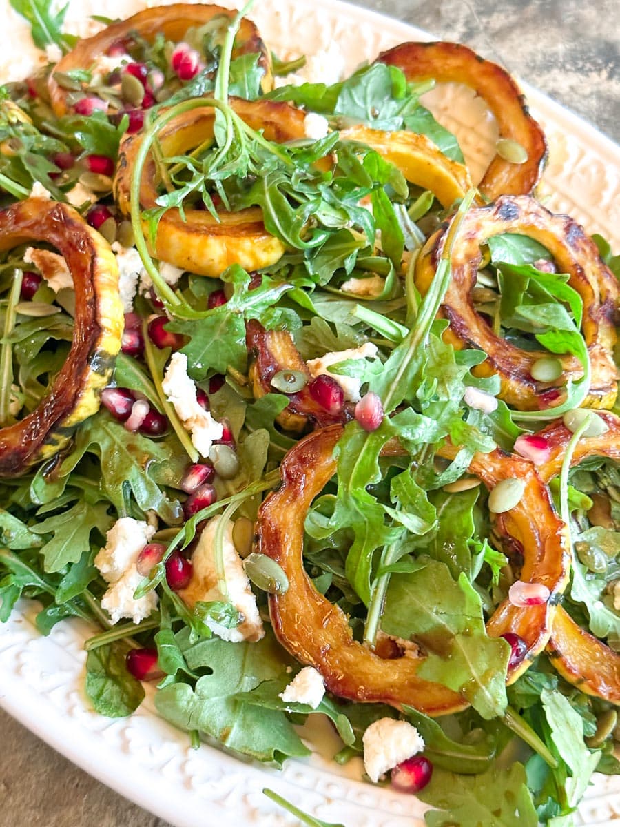 closeup of Low FODMAP Roasted Delicata Squash & Arugula Salad with Goat Cheese and Pomegranate on a white oval platter.