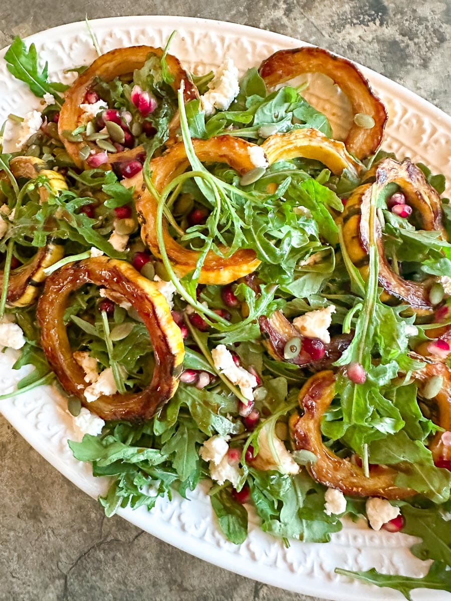 vertical image of Low FODMAP Roasted Delicata Squash & Arugula Salad with Goat Cheese and Pomegranate on a white oval platter.