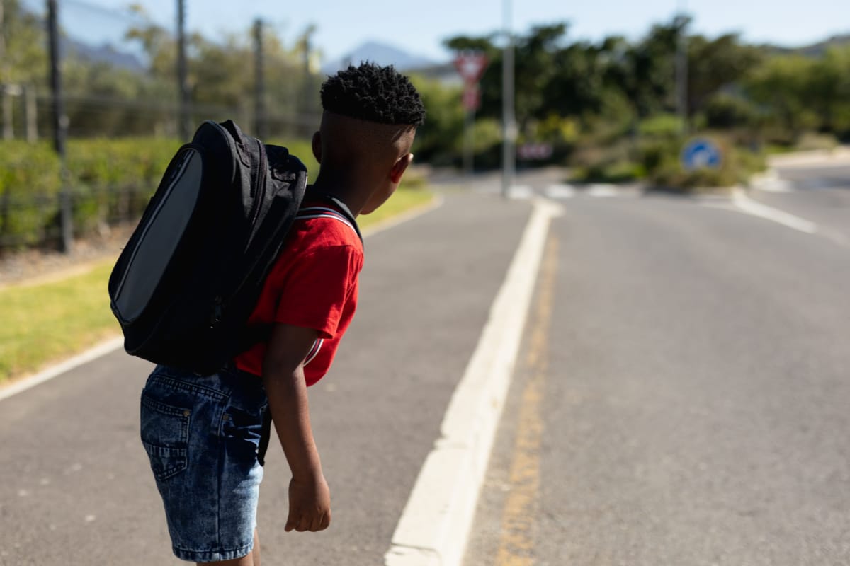 Side view of an African American schoolboy wearing shorts and a t shirt and carrying a rucksack, looking away from camera for traffic while waiting to cross the road on the way to elementary school.