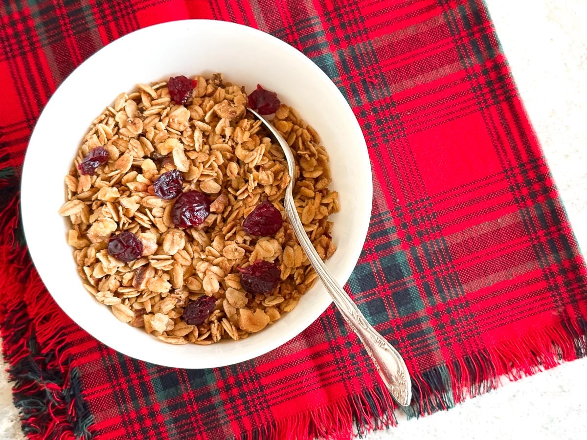 Low FODMAP Maple Walnut Granola on white bowl with dried cranberries on red plaid.