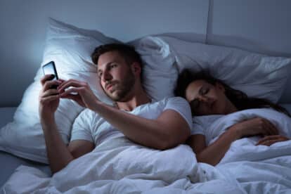Young Man Using Cellphone While Her Wife Sitting On Bed At Night.