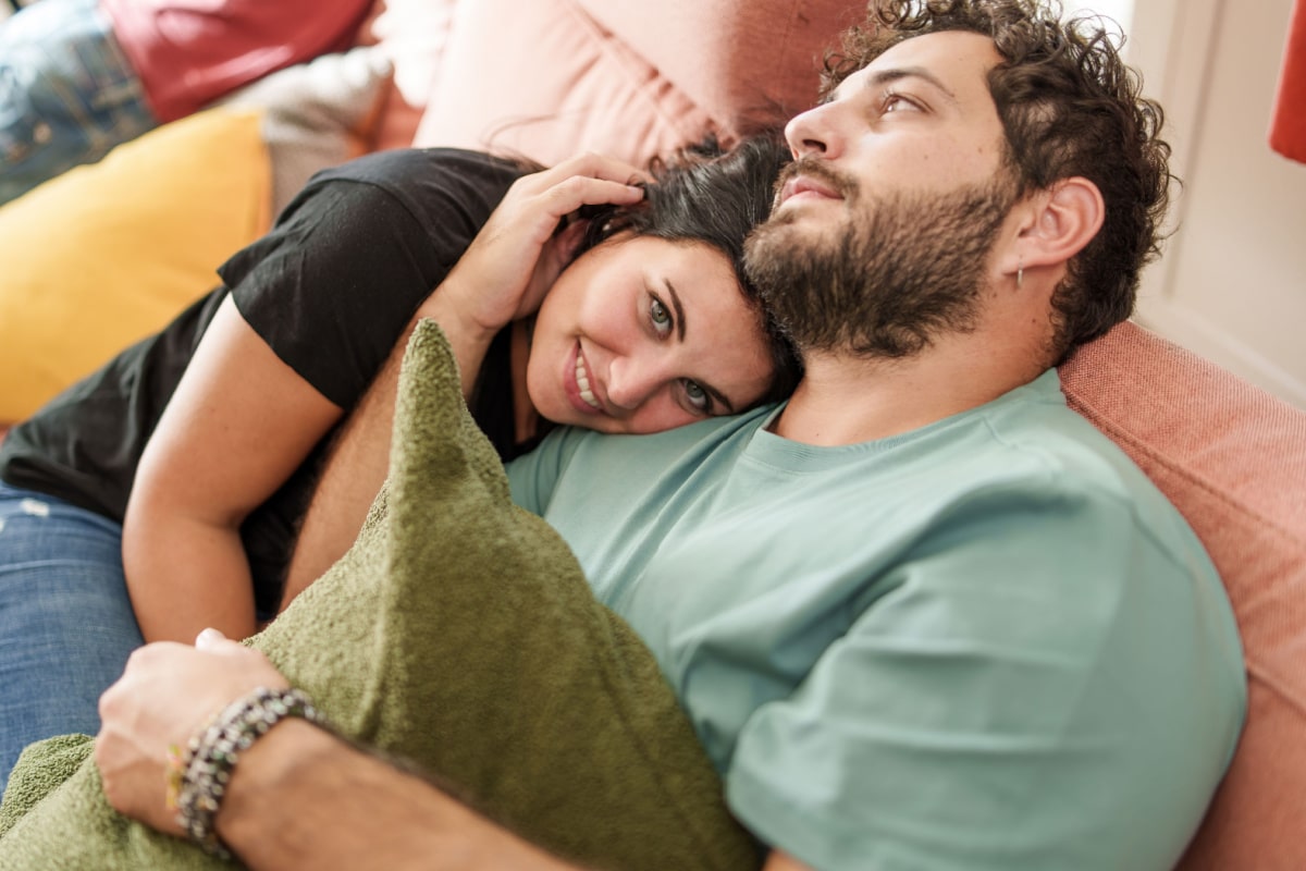 A woman with a warm smile leans on her partner's chest, sharing an intimate and relaxed moment together on a couch, surrounded by soft cushions, showcasing their deep connection.