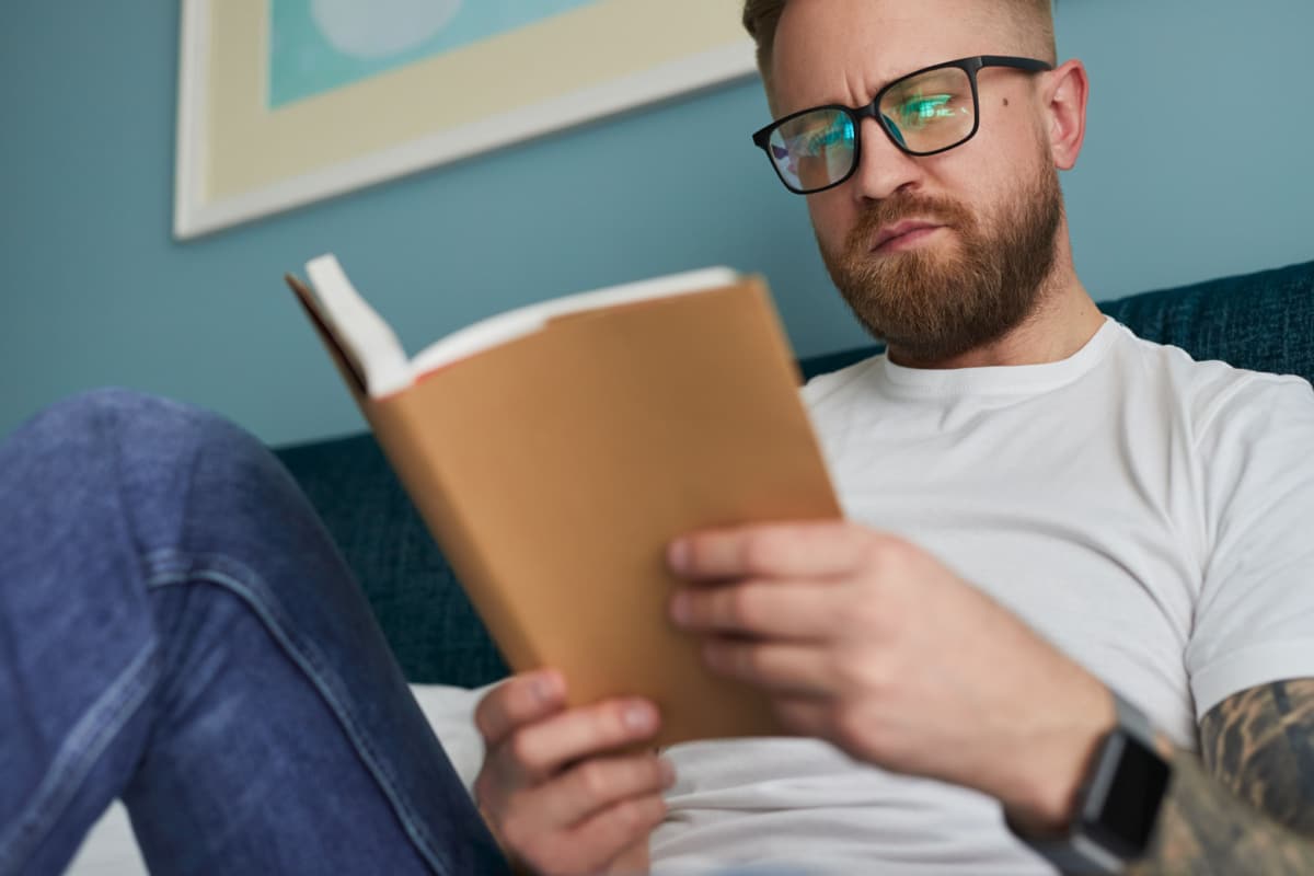 Low angle of focused adult bearded male in eyeglasses reading interesting book while spending free time at home. Intelligent man. Intellect. 