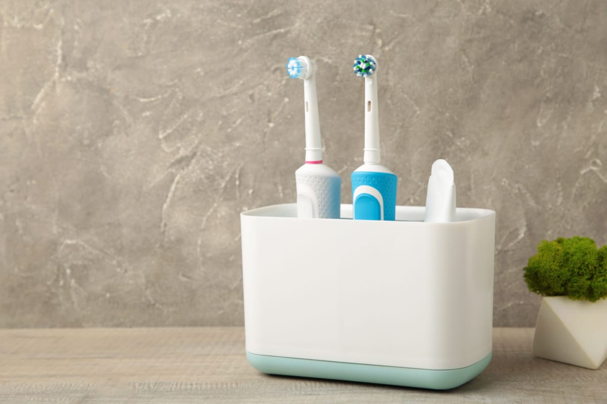 Holder with modern electric toothbrushes and plant on grey background.