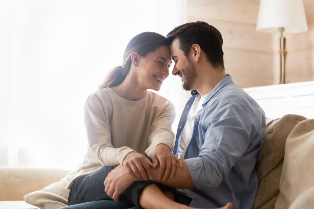 Happy together. Smiling newlyweds cuddling on couch at living room of rented purchased flat, caring husband hugging holding on knees beloved wife, young couple is glad to start new independent life.