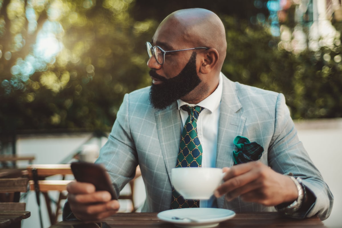 A portrait of a handsome dapper bald mature black man entrepreneur with a well-groomed beard and in a formal suit with a necktie, sitting in a street restaurant and drinking tea during a coffee break.