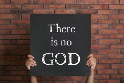 Atheism. Man holding sign with text There Is No God near brick wall, closeup